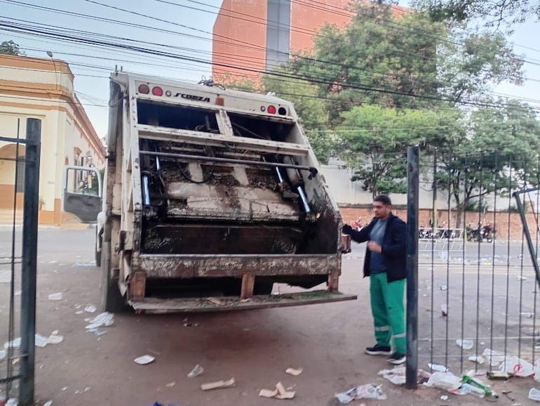 Hombre en pantalones verdes y chaqueta negra supervisa camión de basura en un suelo cubierto de desechos en un entorno urbano.