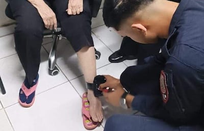 Mujer en ropa oscura y pantuflas rosas observa al hombre con uniforme azul oscuro mientras le coloca un dispositivo de monitoreo en el tobillo.
