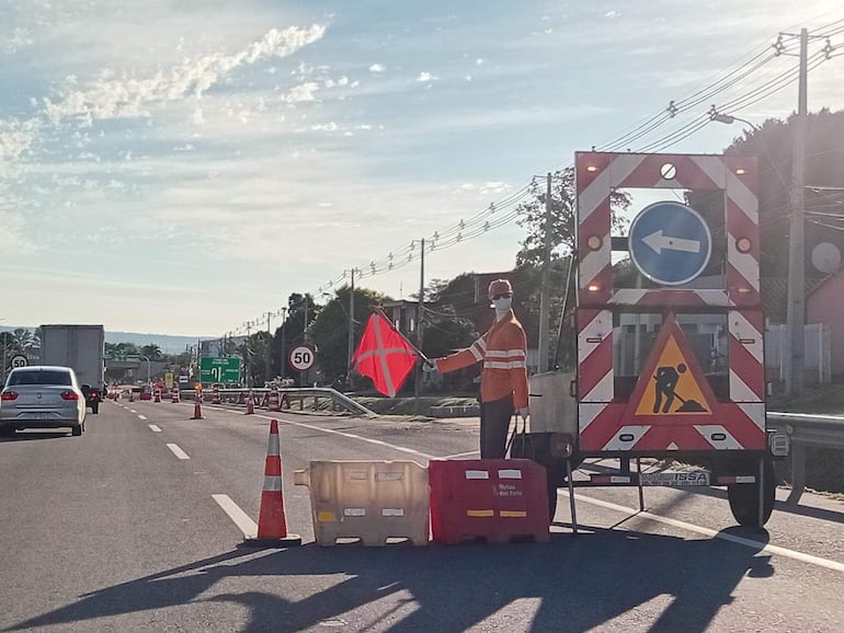 Trabajador vial con chaqueta de seguridad anaranjada y casco rojo dirige el tráfico en la carretera.