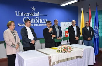 Mujer con abrigo claro y sacerdote reflexionan mientras orador principal con micrófono habla, rodeados de banderas diocesanas.