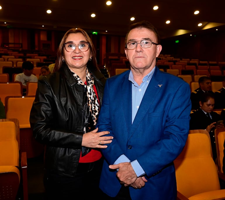 Mujer con chaqueta de cuero negra y bufanda de leopardo sonríe, mientras hombre con blazer azul observa serio en un auditorio.