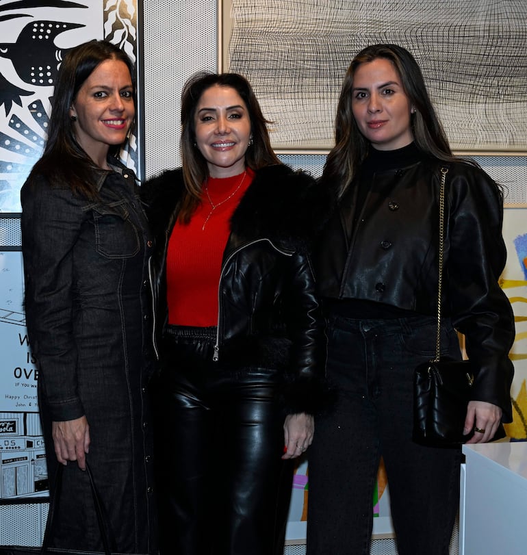 Tres mujeres sonrientes, una en blusa roja y abrigo de piel negra, posan en un interior decorado con obras de arte.