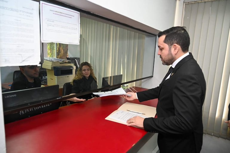 Luis Bello, vestido formalmente, entrega documentos a una mujer de cabello rizado y vestido negro en un ambiente de oficina.