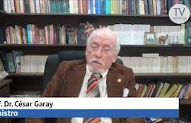 César Garay Zuccolillo, hombre canoso en saco marrón, se sienta frente a estantería de libros, con corbata de rayas naranja y negro.
