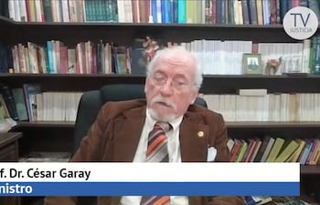 César Garay Zuccolillo, hombre canoso en saco marrón, se sienta frente a estantería de libros, con corbata de rayas naranja y negro.