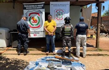 Hombre esposado en camiseta amarilla, custodiado por policías en uniforme. Manta con armas y municiones en el suelo.