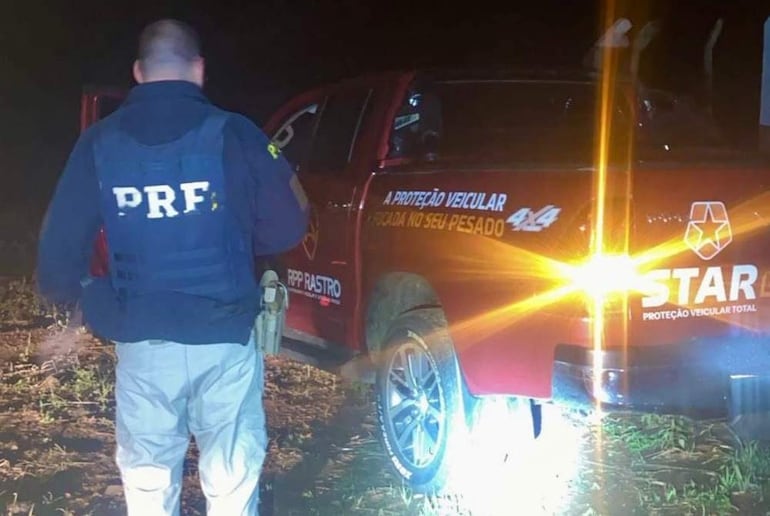 Hombre con chaleco PRF de espaldas frente a camioneta roja con luces encendidas en ambiente nocturno.