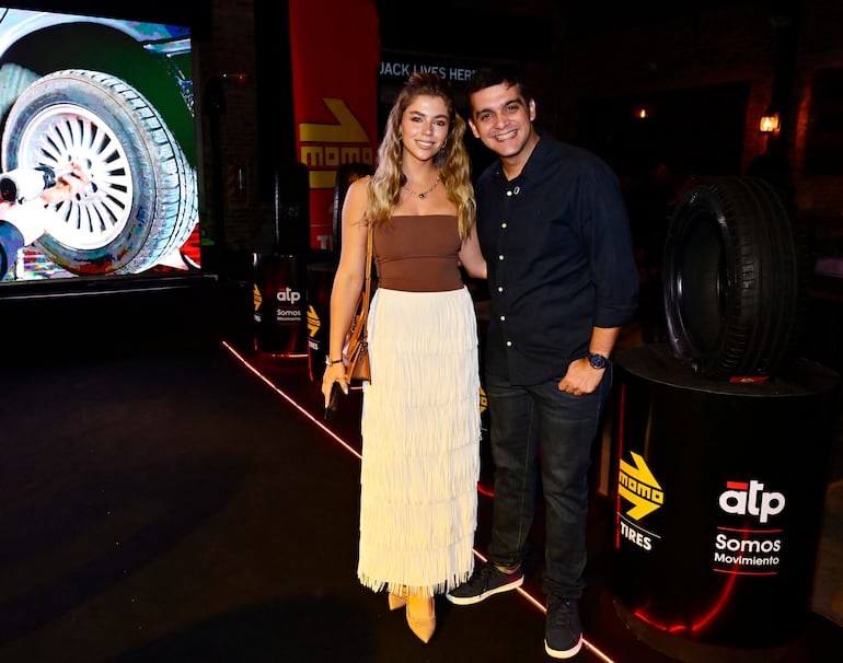 Hombre en camisa oscura y jeans junto a mujer en top marrón y falda blanca con flecos, ambos sonrientes en evento de Momo Tires.