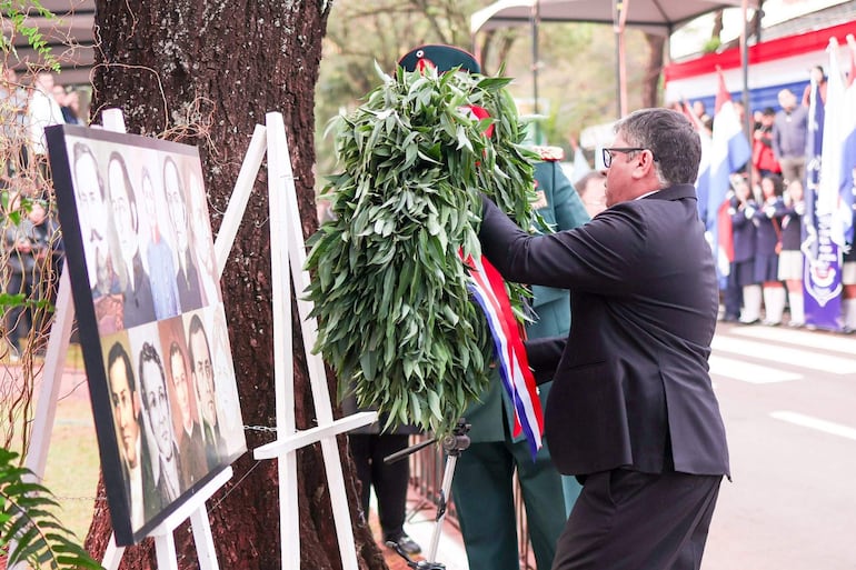 Hombre en traje oscuro sostiene ofrenda de hojas verdes, mientras se aproxima a un soldado en uniforme militar bajo vegetación.