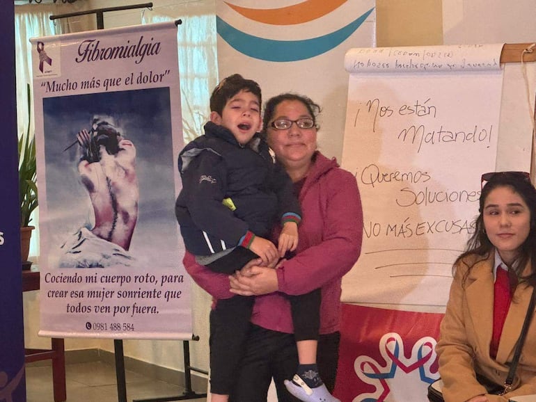 Mujer con chaqueta morada sostiene a un niño con expresión de incomodidad, mientras otra mujer observa con prenda beige.