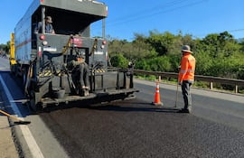 Tres trabajadores en ruta PY01; uno con rastrillo, otro cerca de un camión de asfalto, cielo despejado y vegetación al fondo.