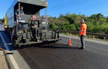 Tres trabajadores en ruta PY01; uno con rastrillo, otro cerca de un camión de asfalto, cielo despejado y vegetación al fondo.
