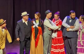 Seis actores en el escenario, con trajes formales y coloridos, interactúan en un ambiente teatral oscuro.