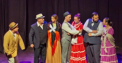 Seis actores en el escenario, con trajes formales y coloridos, interactúan en un ambiente teatral oscuro.