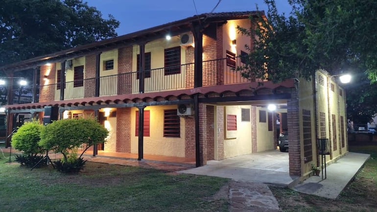 Edificio de dos pisos iluminado, con ladrillos y paredes amarillas, rodeado de césped y arbustos, sin personas visibles.