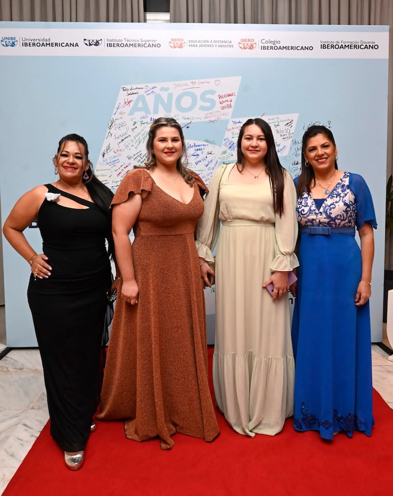 Cuatro mujeres elegantes sonríen en un evento, visten vestidos de diversos colores, en un ambiente festivo con fondo decorativo.