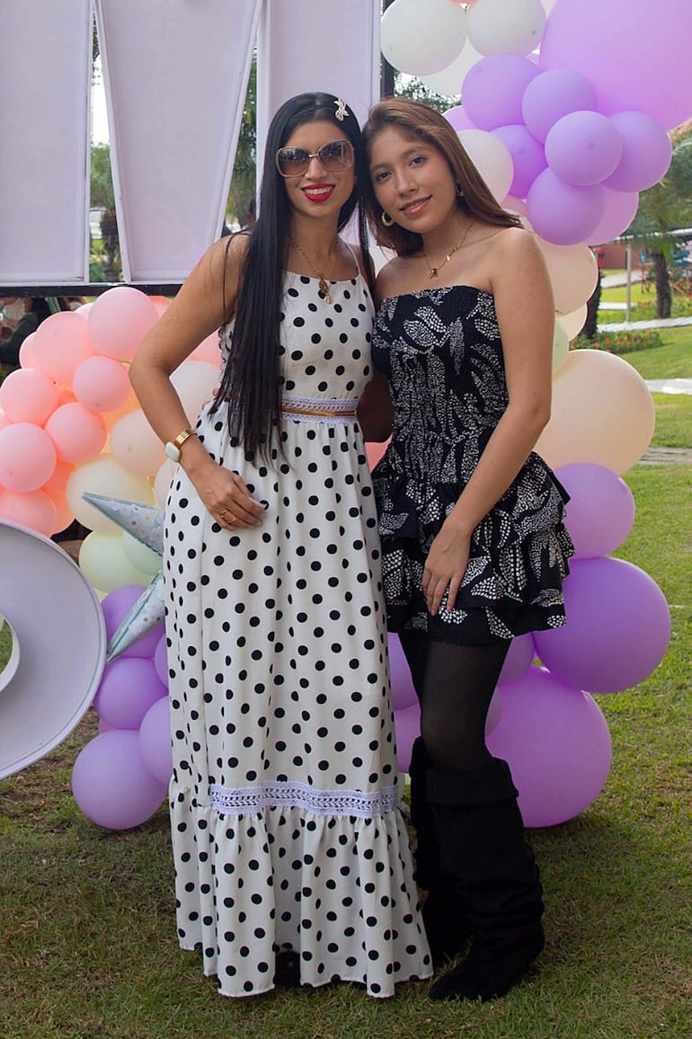 Dos mujeres sonrientes, una en vestido blanco con lunares, otra en vestido floral negro, entre globos en un jardín al aire libre.