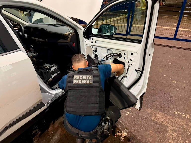 Oficial de la Receita Federal, vestido con chaleco táctico, inspecciona un auto blanco con interior expuesto.