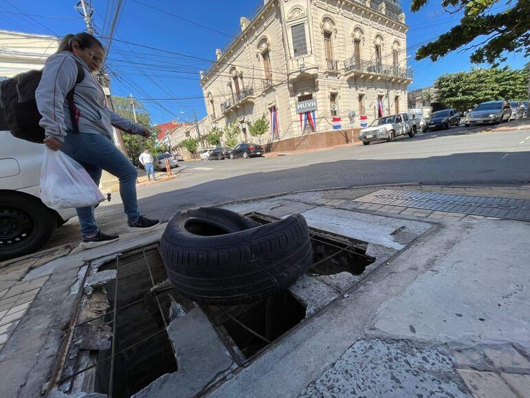 Mujer con mochila y bolsa de compras camina sobre acera en Asunción, evitando neumático. Edificio clásico y cielo despejado al fondo.