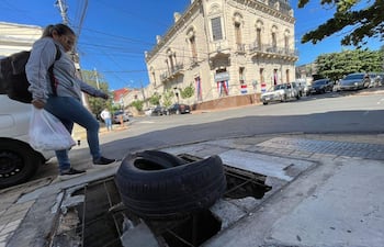 Mujer con mochila y bolsa de compras camina sobre acera en Asunción, evitando neumático. Edificio clásico y cielo despejado al fondo.