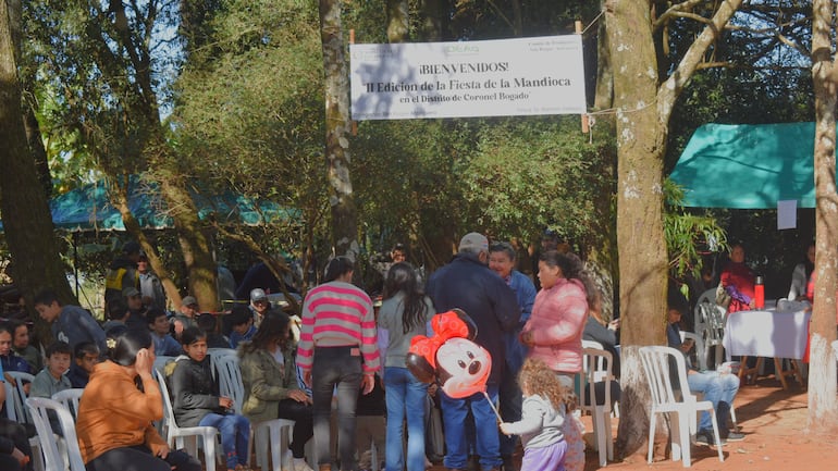 Varias personas conversan y juegan en un evento al aire libre rodeado de árboles y un cartel de bienvenida.