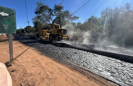 Trabajador con chaleco naranja observa máquina amarilla aplicando asfalto en carretera, rodeado de vegetación densa.