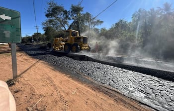 Trabajador con chaleco naranja observa máquina amarilla aplicando asfalto en carretera, rodeado de vegetación densa.