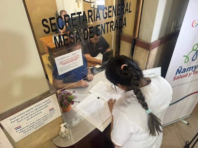Mujer con camiseta blanca revisa documentos en escritorio, mientras dos hombres conversan tras vidrio en oficina iluminada.