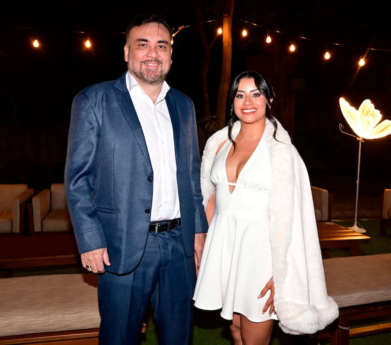 Hombre de traje oscuro y mujer en vestido blanco, ambos sonrientes en un evento al aire libre con luces decorativas.