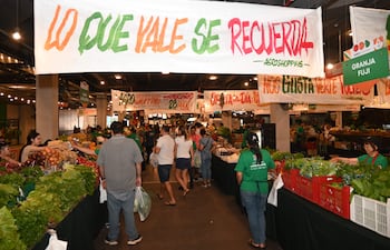 Personas compran en Agroshopping, rodeadas de frutas y verduras, con un ambiente festivo y trabajadores en camisetas verdes.