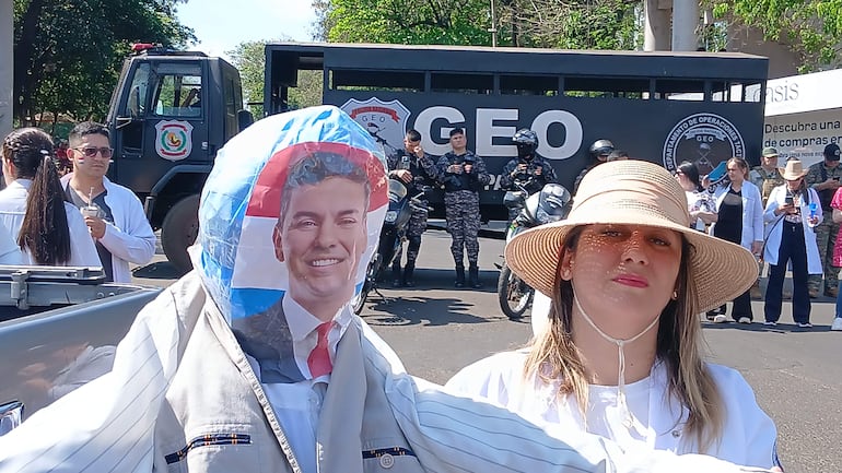 Mujer con sombrero de ala ancha y blusa blanca, junto a hombre que sostiene muñeco de rostro sonriente en manifestación frente a la policía.