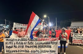 Mujer en el centro sostiene una bandera paraguaya mientras otros manifestantes levantan carteles. Ambiente nocturno de protesta.