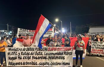 Mujer en el centro sostiene una bandera paraguaya mientras otros manifestantes levantan carteles. Ambiente nocturno de protesta.