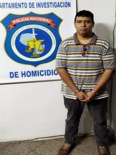 Hombre de pie con camiseta de rayas, esposado frente al mural del Departamento de Investigación de Homicidios, en ambiente sobrio.