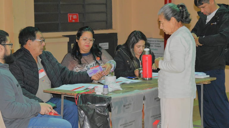 Cinco personas en un centro de votación, tres sentadas ante una mesa con documentos y dos de pie, una anciana sosteniendo un objeto.