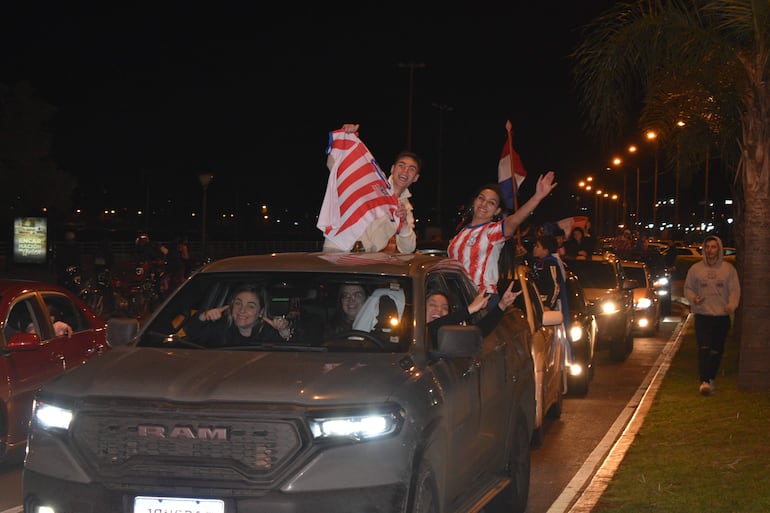 Cinco hinchas de pie sobre una camioneta, vistiendo camisetas rojas y blancas, celebran con alegría en una noche festiva.