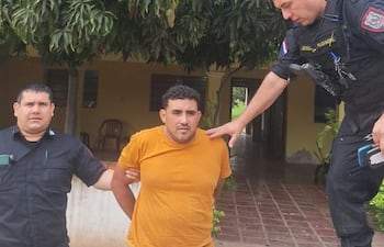 Hombre en camiseta amarilla bajo custodia de policías, rodeado de vegetación en un ambiente exterior, mirada neutral y postura controlada.
