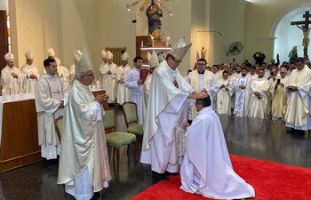 Sacerdote arrodillado recibiendo imposición de manos vestido con túnica blanca en ceremonia solemne, rodeado de otros sacerdotes.