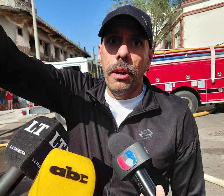 Héctor Vergara con bigote y gorra oscura, con camiseta blanca y chaqueta negra, rodeado de micrófonos en un ambiente de emergencia.