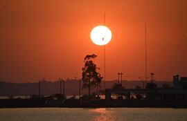 Atardecer con sol brillante sobre cuerpo de agua, siluetas de árboles y postes en un ambiente sereno.