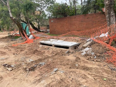 Zona de obras con tierra removida y escombros, además de un par de árboles y una malla de obra naranja iluminada por el sol.