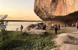 Dieciséis personas distribuidas en un acantilado, contemplando un río con atardecer en tonos cálidos, algunas tomando fotografías.