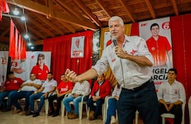 Arnoldo Wiens en camisa blanca, hablando con energía frente a una audiencia de mujeres y hombres, rodeado de carteles políticos.