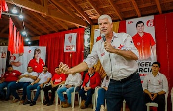 Arnoldo Wiens en camisa blanca, hablando con energía frente a una audiencia de mujeres y hombres, rodeado de carteles políticos.
