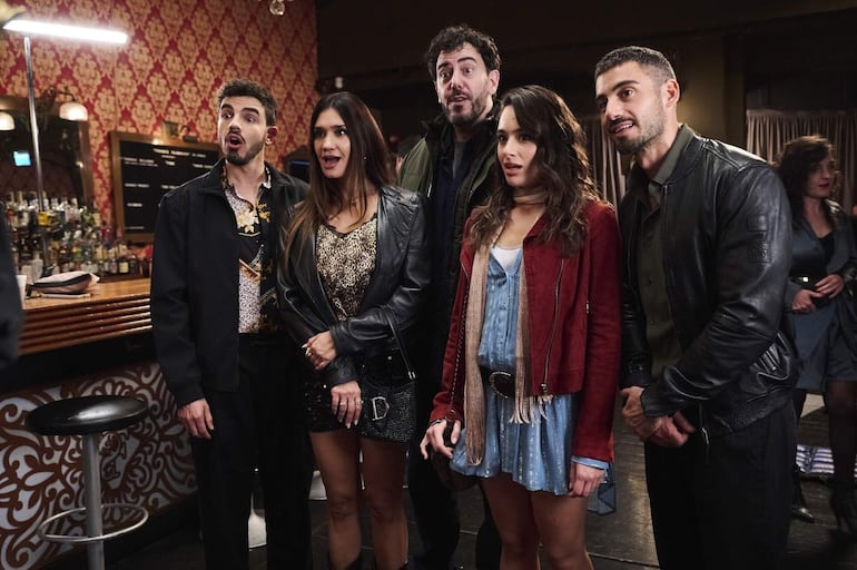 Grupo de cinco personas en bar. Hombre con chaqueta oscura, mujer con blusa de animal print, hombre con abrigo oscuro, mujer con bufanda y hombre con chaqueta negra.