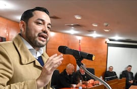 Luis Bello con abrigo claro gesticulando frente a un micrófono, rodeado de hombres sentados en una mesa atenta.