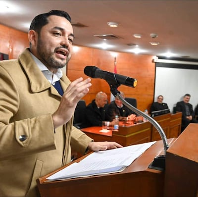 Luis Bello con abrigo claro gesticulando frente a un micrófono, rodeado de hombres sentados en una mesa atenta.