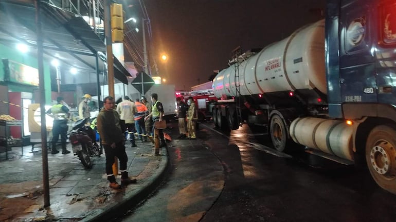 Cinco personas en la escena de un accidente: uniformes de bomberos y civiles, luces de emergencia, un camión tanque en el fondo.