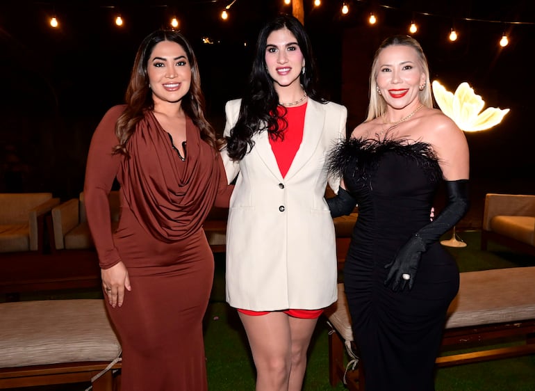 Tres mujeres sonrientes: una en vestido marrón, otra en abrigo blanco y vestido rojo, y la última en vestido negro con plumas.