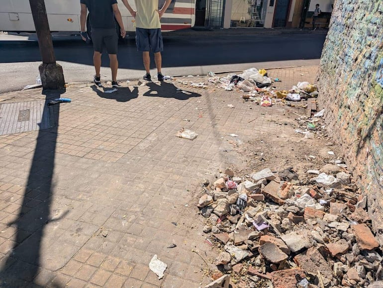 Hombres de espaldas, uno con camiseta oscura y otro con camiseta clara, frente a una vereda cubierta de basura y escombros.